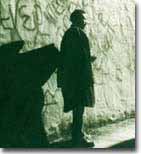 Full length image of Alexander stood against wall with graffiti. He is turned away from the camera and wearing a large jacket. He is casting a shadow on the wall.  