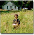 Image of Bobbie West stood in a field. The grasses are as high as her neck. There is a small, white building in the background.