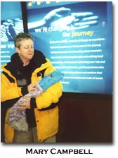 Campbell poses in front of an informative sign wearing a large jacket while cradling a bundle of material