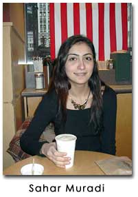 Muradi sits at a table holding a cup and smiles. The red and whites stripes of a U.S. flag hangs behind her