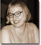 Sepia headshot of Kathy Lou Schultz. She is wearing black glasses and a floral dress. 