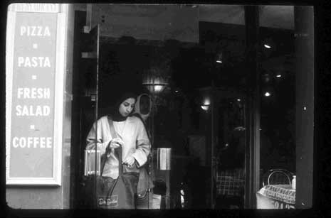 A black and white photograph of a woman wearing a head covering standing in a storefront window